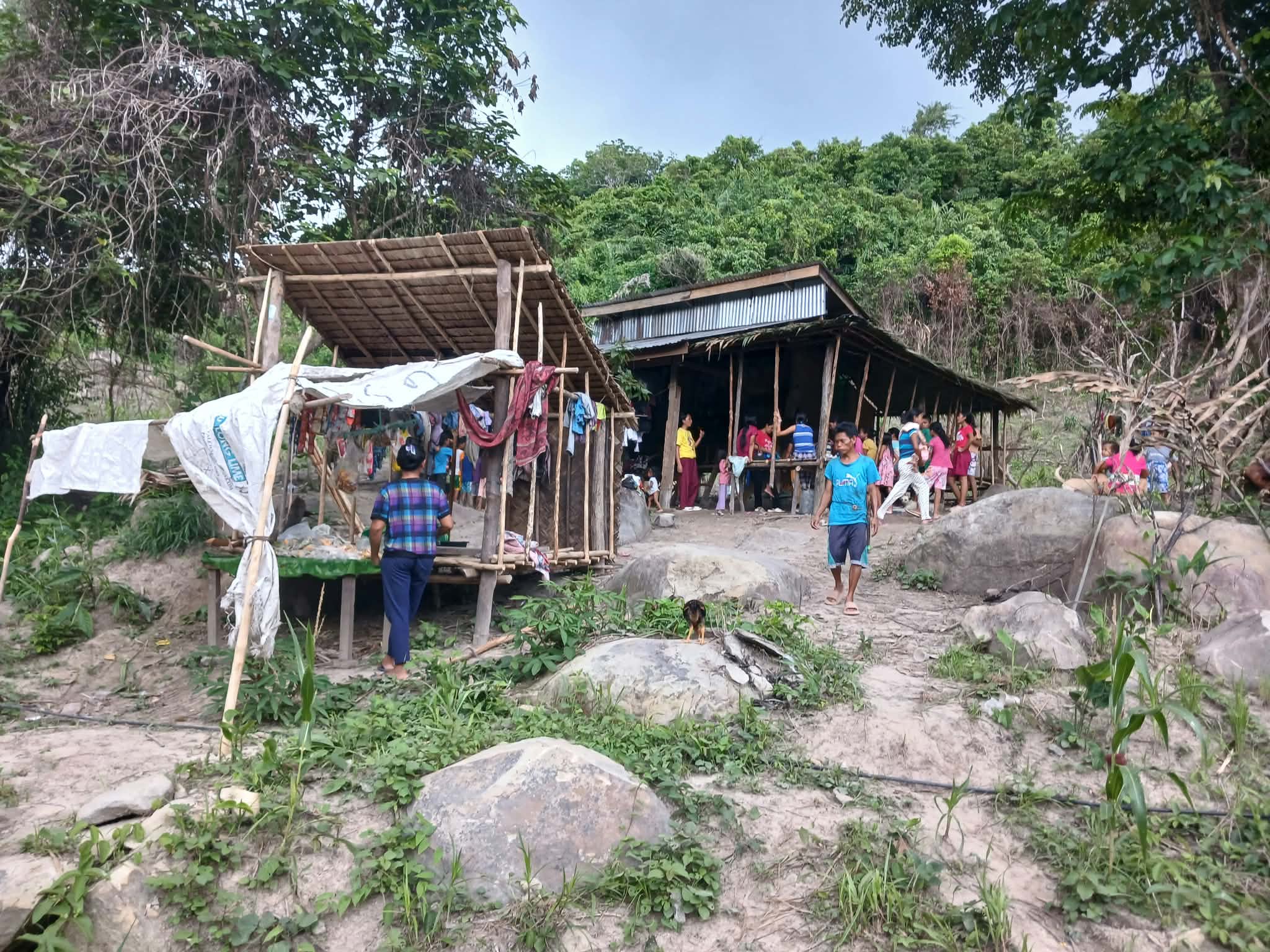 Community members gathered near simple rural structures in a rocky hillside setting